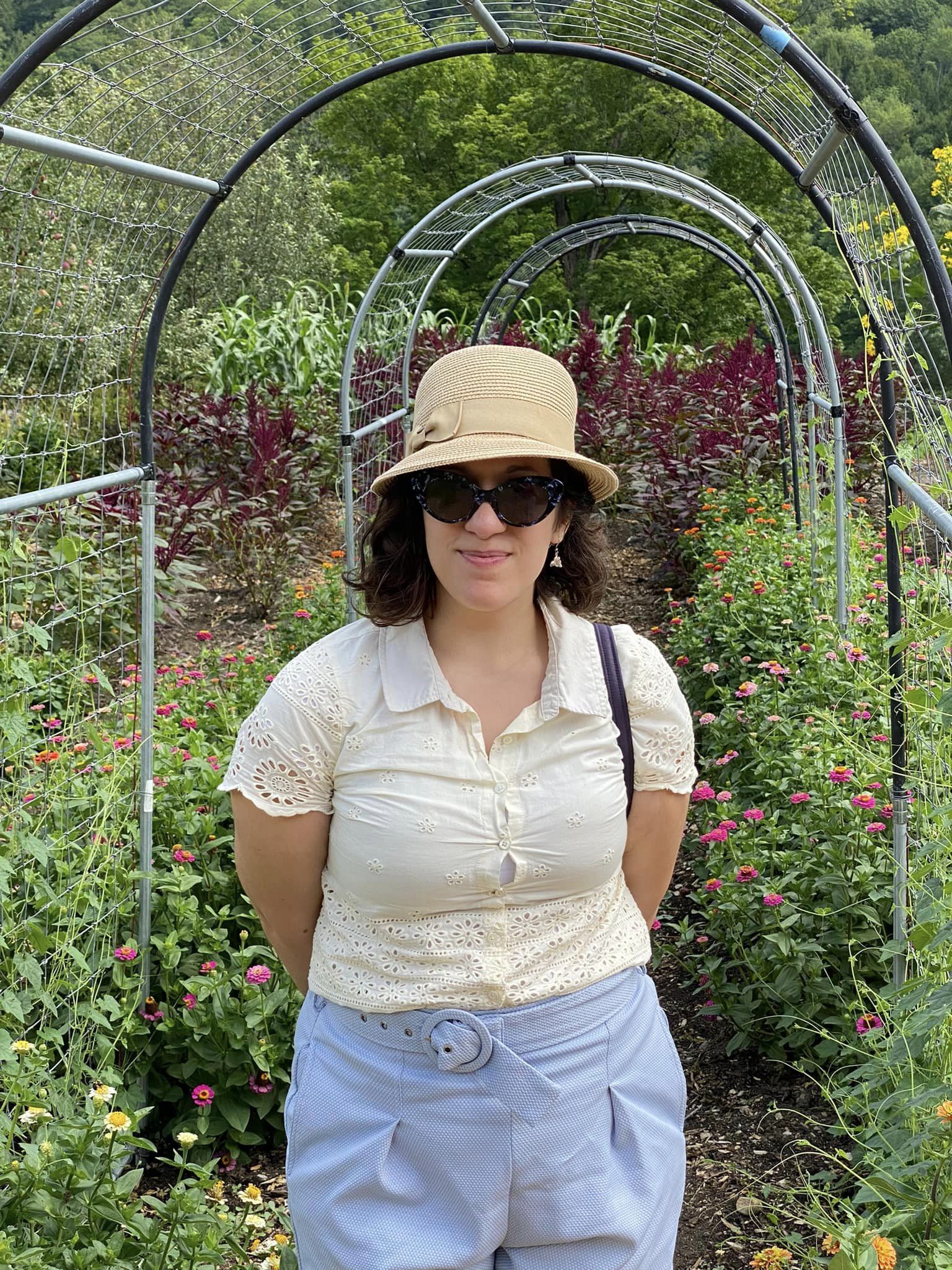 Smiling woman with sunglasses, woven hat and short brown curly hair, under an arch in a lush garden